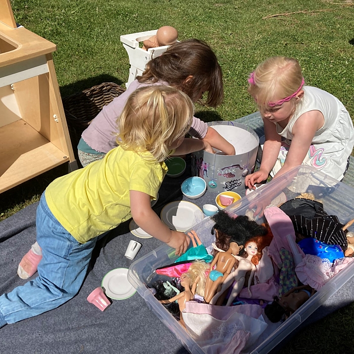 Billede af tre børn, der leger med dukker på et tæppe på legepladsen.