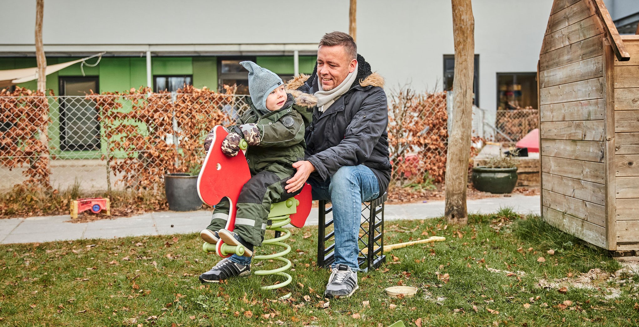 Billede af en dreng, der vipper på legepladsen mens han kigger bagud til en voksen, som smilende sidder og passer på, han ikke falder ned.